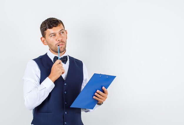 Waiter holding clipboard and pencil in shirt, vest and looking pensive , front view.