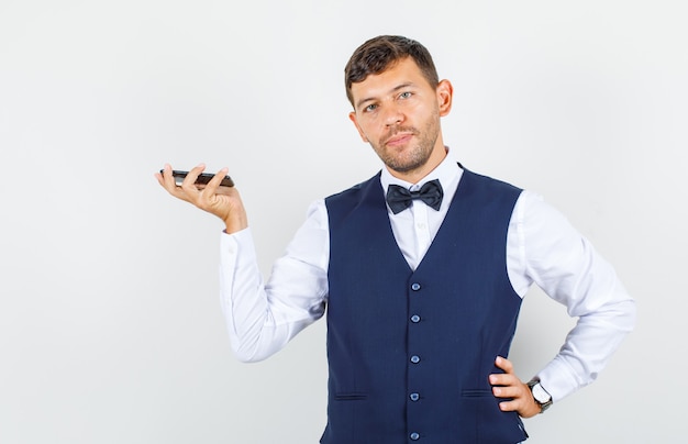 Waiter holding mobile phone in shirt, vest and looking confident , front view.