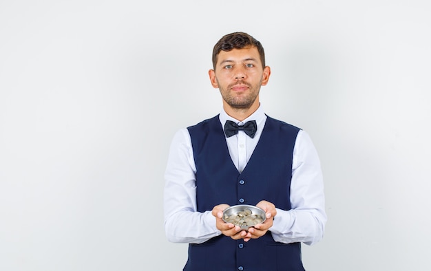 Waiter holding pile of coins in shirt, vest and looking confident , front view.