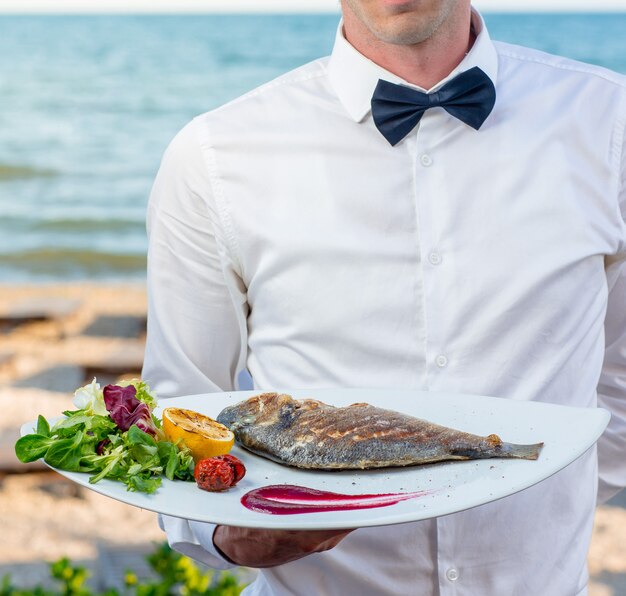 Официант держит тарелку жареной рыбы с жареным лимоном, помидорами, свежим шпинатом, салатом