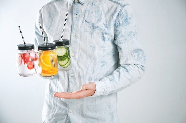 Waiter holds three cold refreshment beverages from strawberry, orange, lime, mint, cucumber, ice and sparkling water in rustic jars with drinking straws inside