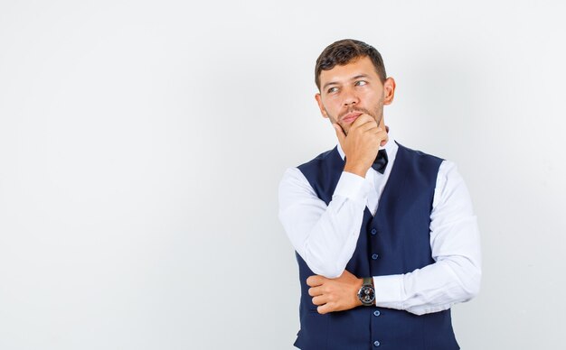 Waiter looking aside with hand on chin in shirt, vest and looking pensive , front view.