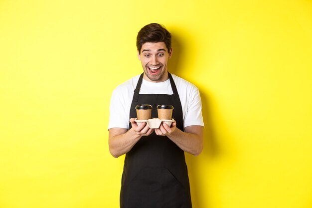 Waiter looking excited at two cups of takeaway coffee, wearing black apron, standing over yellow background