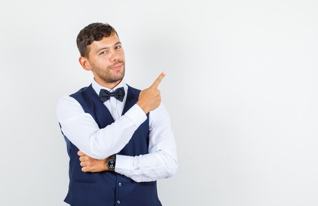 Waiter pointing away and smiling in shirt, vest and looking confident. front view.