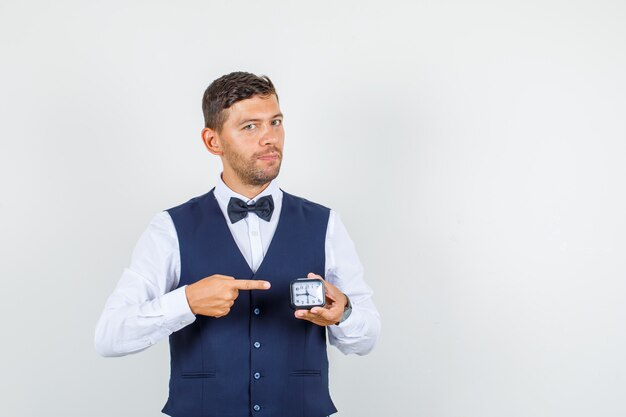 Waiter pointing at clock in shirt, vest and looking confident , front view.