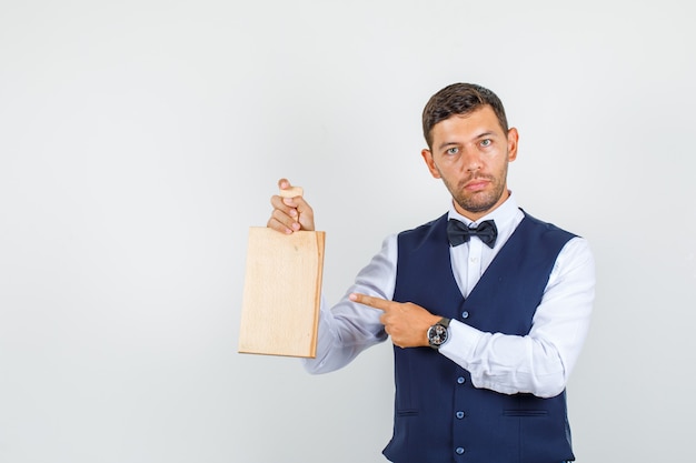 Waiter pointing at cutting board in shirt, vest and looking serious. front view.