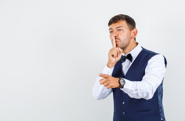 Waiter pointing finger away with silence gesture in shirt, vest and looking careful , front view.