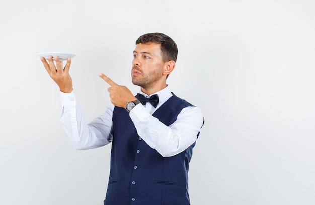 Free photo waiter pointing finger at empty plate in shirt, vest and looking serious , front view.