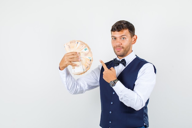 Waiter pointing finger at euro banknotes in shirt, vest and looking cheerful. front view.