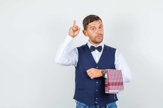 Waiter pointing finger up in shirt, vest, jeans and looking serious , front view.