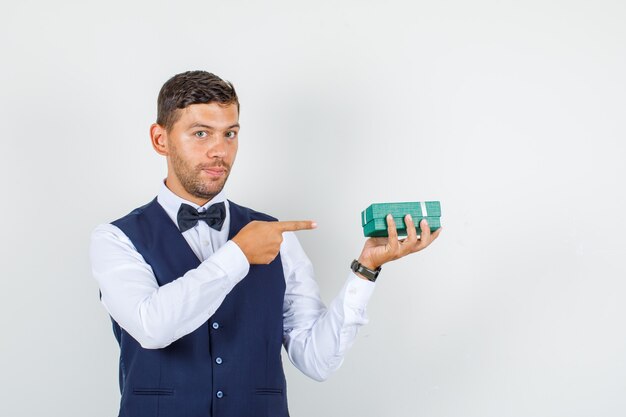 Waiter pointing at gift box in shirt, vest and looking confident. front view.