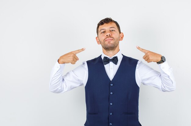 Waiter pointing index fingers at himself in shirt, vest and looking confident , front view.