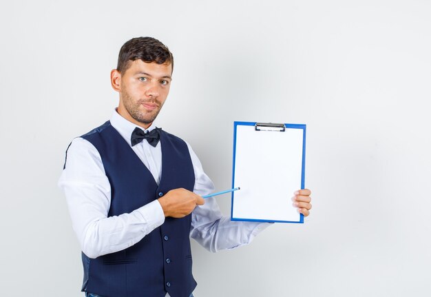 Waiter pointing pencil at clipboard in shirt, vest , front view.