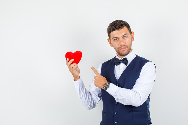 Waiter pointing at red heart in shirt, vest and looking confident. front view.