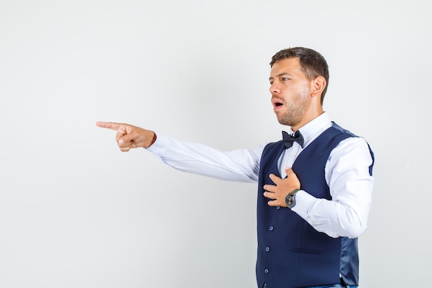 Waiter pointing to side in shirt, vest, bow tie and looking disappointed. front view.