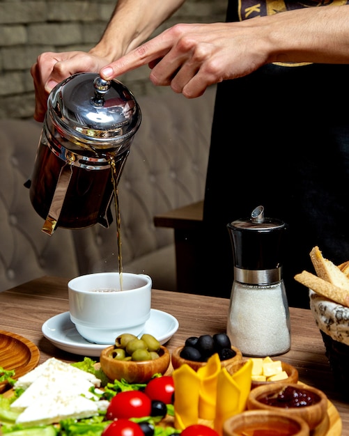 Официант наливает черный чай из французской прессы в стол для завтрака