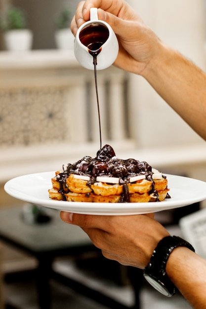 Waiter pours chocolate sauce on waffle with fruits