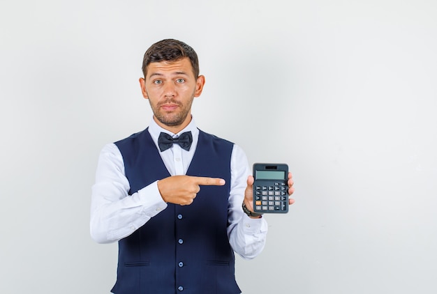 Waiter in shirt, vest, bow tie, pointing at calculator , front view.