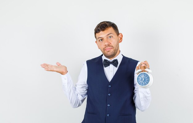 Waiter in shirt, vest holding alarm clock with gesture and looking helpless , front view.