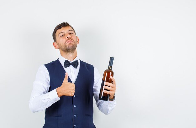 Официант в рубашке, жилете держит бутылку алкоголя с поднятым большим пальцем и гордым видом, вид спереди.