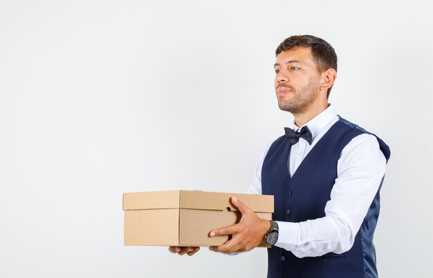 Waiter in shirt, vest holding cardboard box and looking positive , front view.