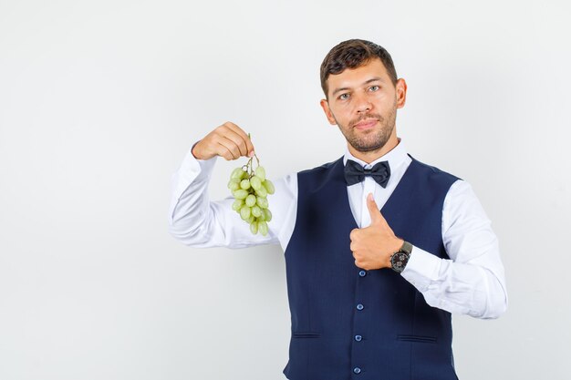 Waiter in shirt, vest, holding grape cluster with thumb up and looking positive , front view.