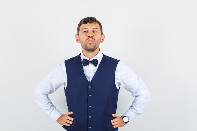 Waiter in shirt, vest holding hands on waist with folded lips , front view.