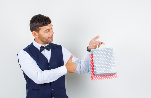 Waiter in shirt, vest holding paper bags with thumb up and looking positive , front view.