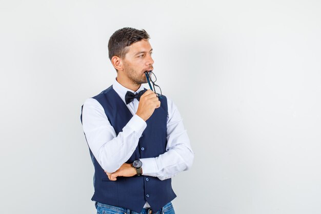 Waiter in shirt, vest, jeans holding glasses near lips and looking dreamy , front view.