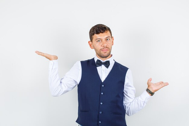 Waiter in shirt, vest raising open palms like holding something and looking serious , front view.