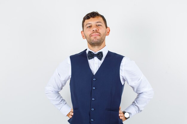 Waiter in shirt, vest standing with hands on waist and looking confident , front view.