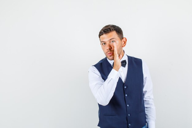 Waiter in shirt, vest telling secret behind hand and looking careful , front view.