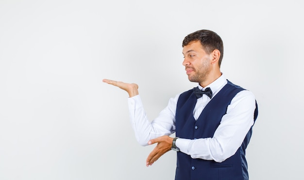 Waiter spreading palm aside and smiling in shirt, vest front view.