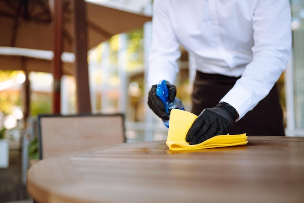 Waiter wearing protective face mask and gloves while disinfecting tables at outdoor cafe