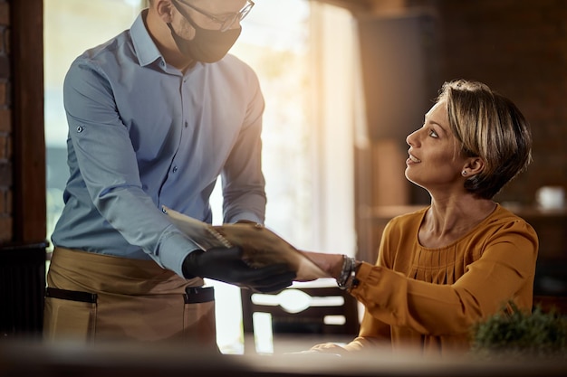 Waiter wearing protective face mask and showing menu to a customer while talking to her in a cafe