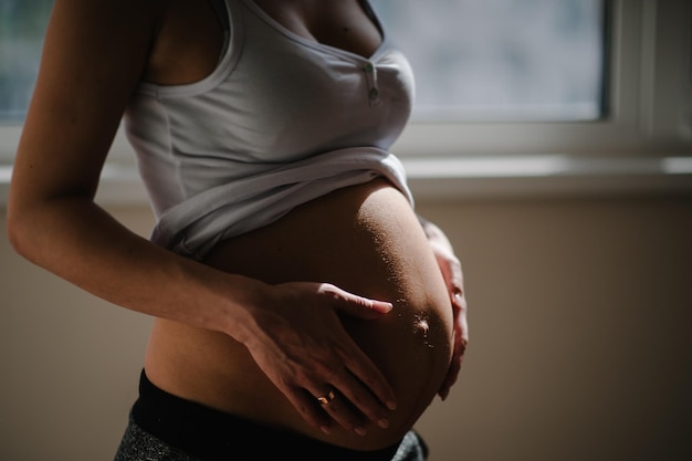 В ожидании ребенка. беременная женщина стоит у окна и обнимает руками круглый живот, живот. крупный план. девять месяцев. детский душ. концепция материнства. вид сбоку.