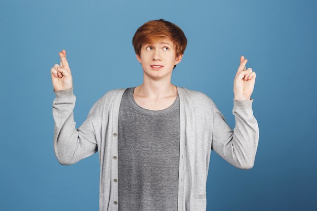 Waiting for exam results, hoping for good mark. Young beautiful male student wit ginger hair in casual outfit holding finger crossed looking aside with worried and tense expression.