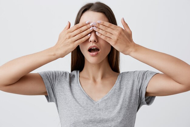 Waiting for exciting moment. Close up of young attractive caucasian woman with long dark hair in casual gray t-shirt clothing eyes with hands, being happy to see birthday present soon.