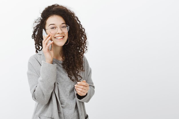 Waiting friend to pick up phone. Confident beautiful curly-haired girl in fashionable coat and glasses, holding smartphone near ear and talking