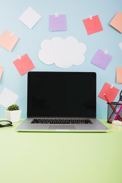 Wall with cloud paper and adhesive notes behind laptop with blank black screen