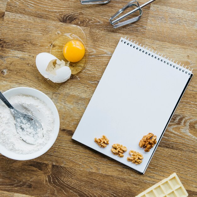 Walnut; spiral notepad; flour; chocolate and whisk on wooden surface