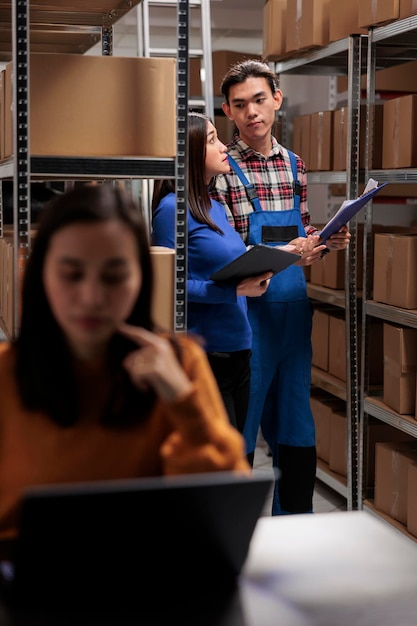 Free photo warehouse asian manager and distribution operator managing stock tracking and looking at cardboard boxes on shelf. storehouse employees searching goods and picking customer order