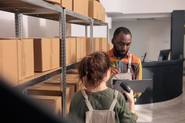 倉庫の従業員がデジタル タブレットのチェックリストを使用して荷物を検索し、発送用の段ボール箱を準備します。オーバーオールを着た起業家が店舗のスキャナーを使用して段ボール箱をスキャン