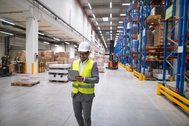 Warehouse manager walking through large storage area and holding tablet while forklift operating in background