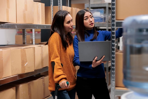 倉庫マネージャーが顧客の注文を準備し、ラップトップでピックチケットを確認します。在庫ソフトウェアを使用しながら棚の上の小包を検索する倉庫の若いアジア人女性従業員