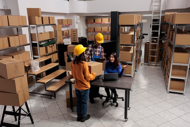 Free photo warehouse package handlers doing inventory control while coworking with logistics manager. asian storehouse employees scanning cardboard box and creating invoice on laptop