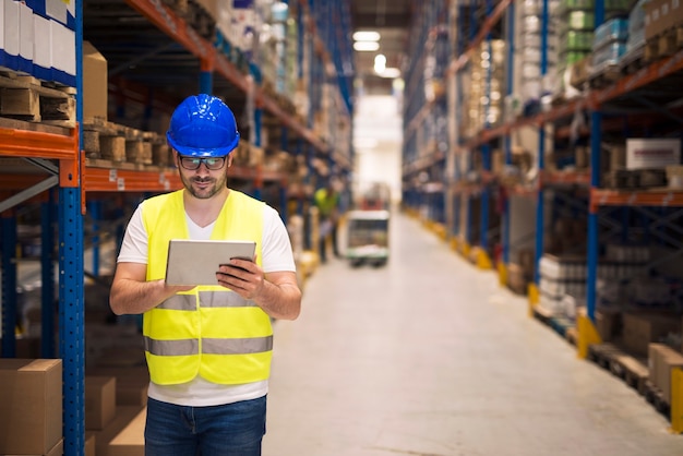 Warehouse worker checking inventory on his tablet while walking in large storage department with shelves and packages in background