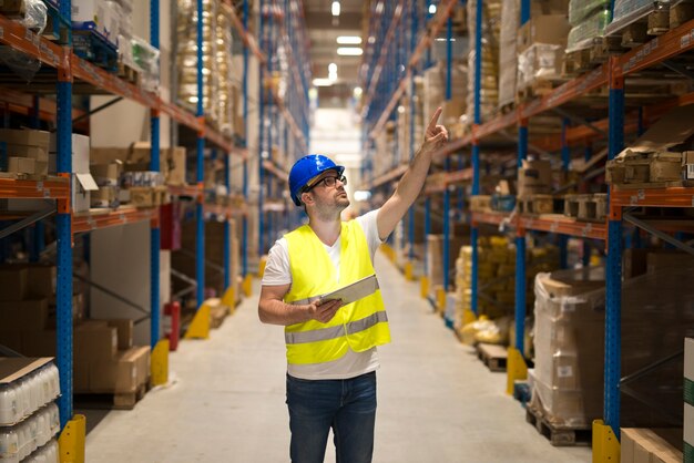 Warehouse worker in protective reflective uniform with hardhat checking inventory and counting product on shelf in large storage area