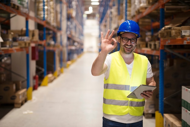 Warehouse worker standing in large storage center and showing OK hand gesture satisfied on delivering goods