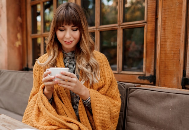Warm cozy portrait of happy dreamy woman with wavy hairs, covered by yellow plaid and holding cup of hot cappuccino. White female chilling on terrace.